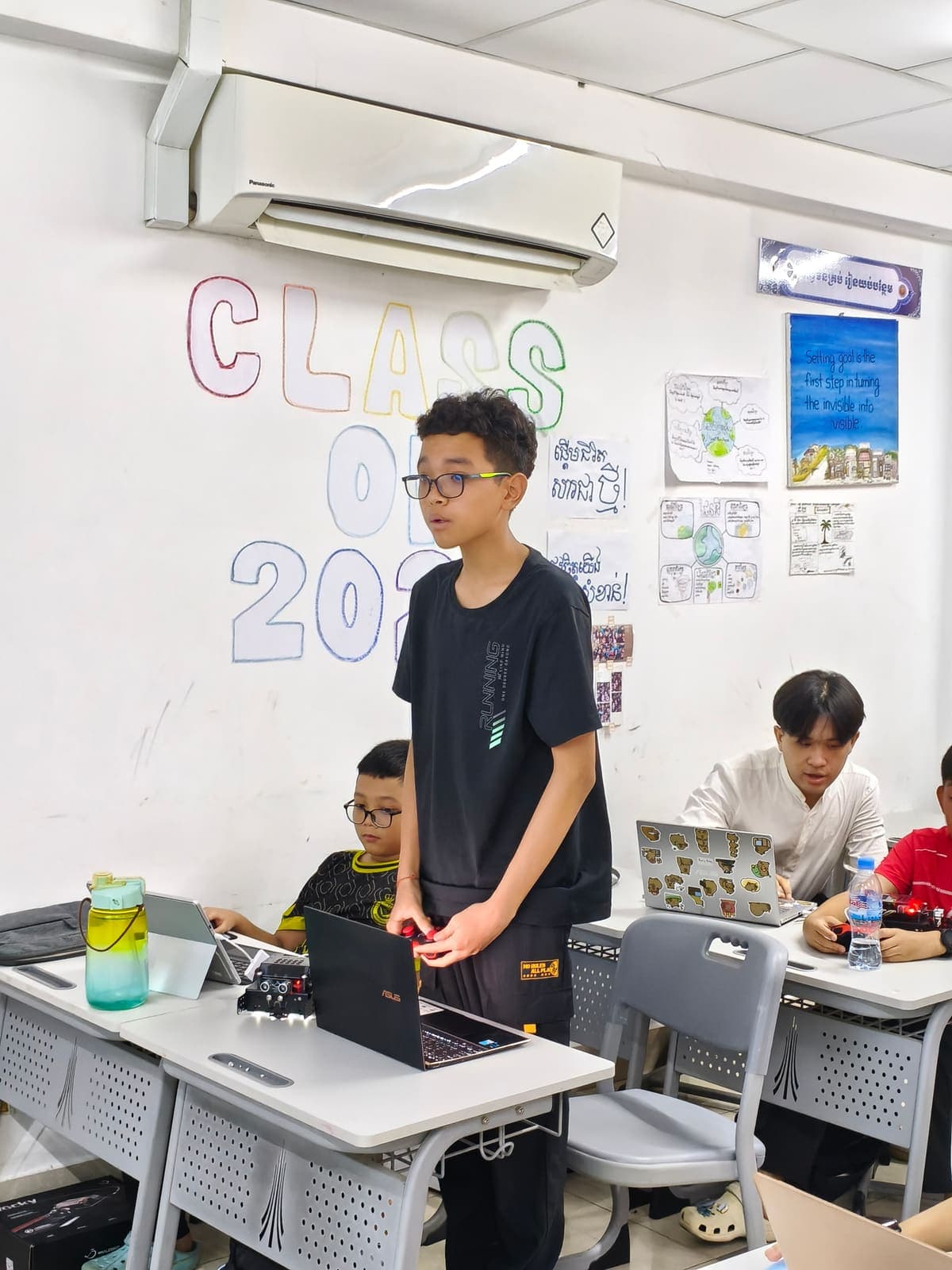 A student standing in a robotics class with a game controller, a robot on the desk in front of a laptop